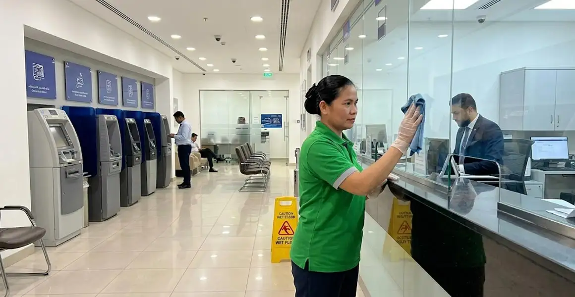A professional commercial cleaner wearing gloves and a green uniform wipes down a glass teller partition with a microfiber cloth inside a modern bank branch lobby in Riyadh, Saudi Arabia.