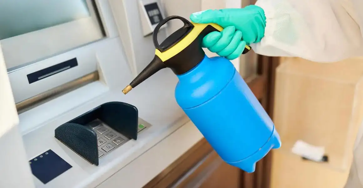 A close-up of a professional cleaning technician wearing green protective gloves using a pressure sprayer to sanitize and disinfect an ATM keypad and screen at a Riyadh bank.
